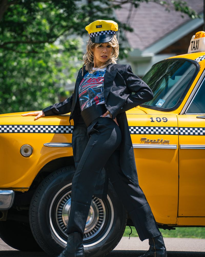 Danae Hays in character with a feathered blonde wig and yellow taxi cap, in a tuxedo jacket and cummerbund, posing against a vintage yellow checker cab.