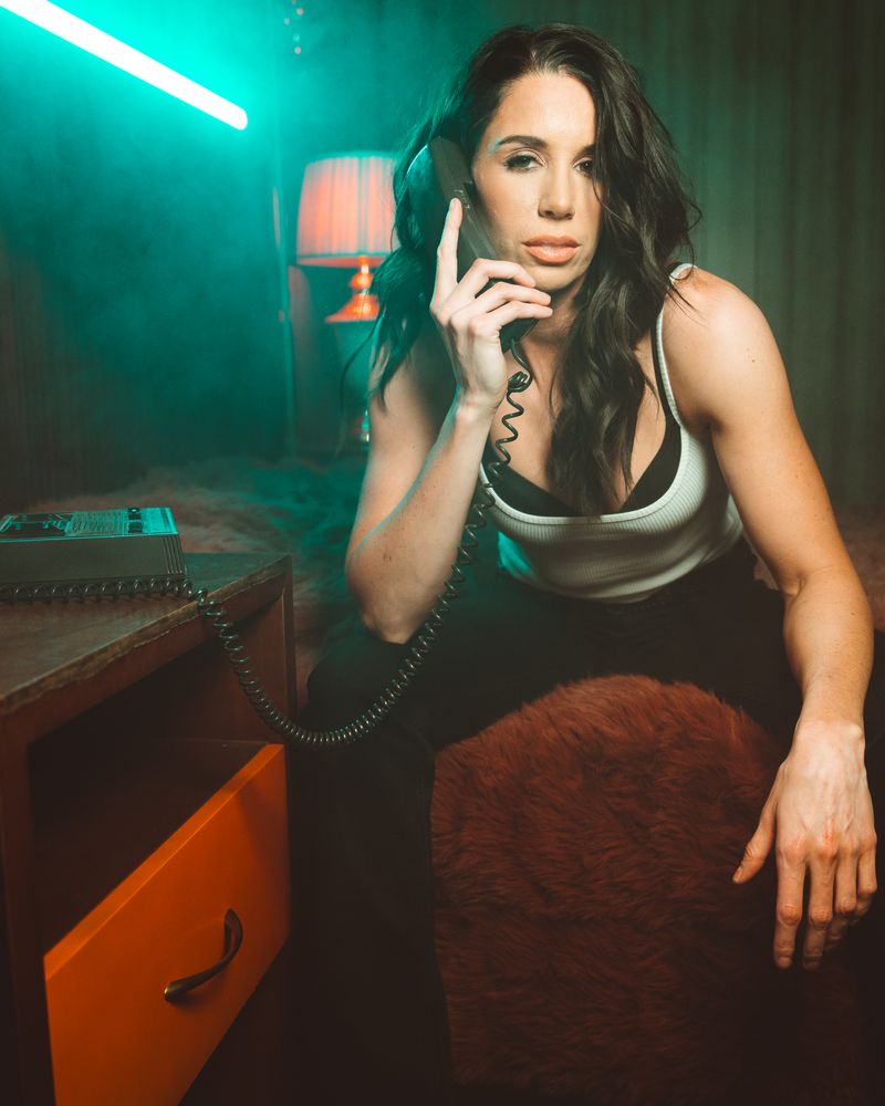 Danae Hays holds a corded phone to her ear with a deadpan look, sitting on a shag-covered bed beside an orange lamp in a smoky, teal-lit room.