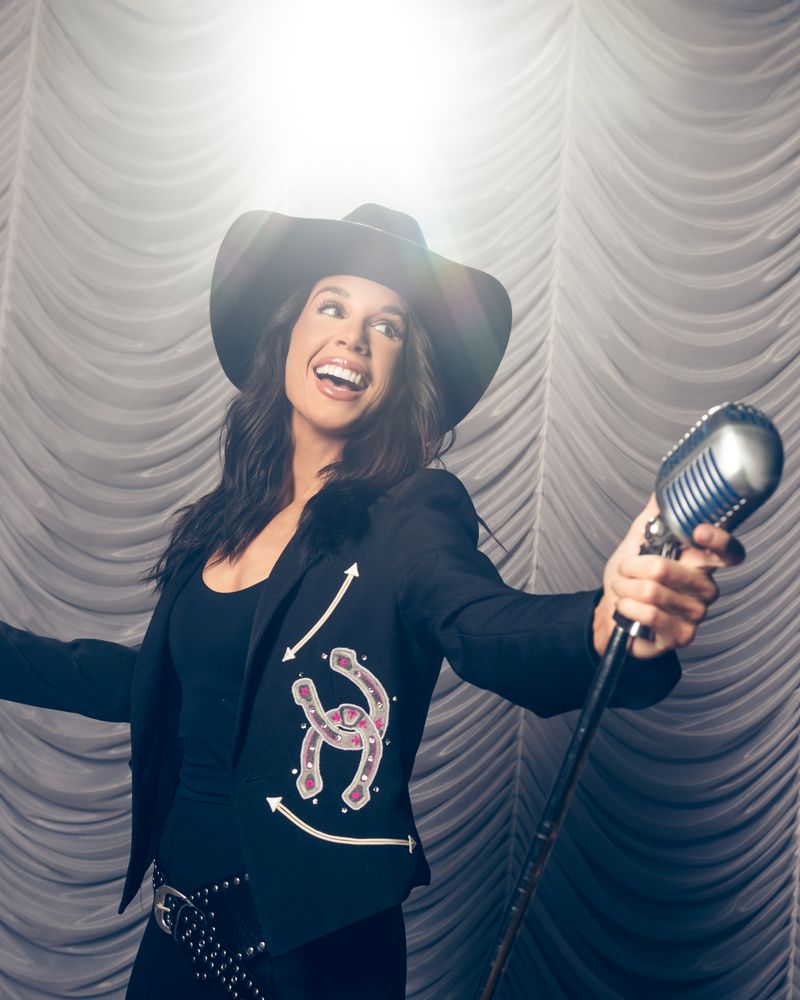 Danae Hays in a black cowboy hat and embroidered horseshoe jacket laughs and holds out a vintage microphone under a bright spotlight, with a draped curtain behind her.