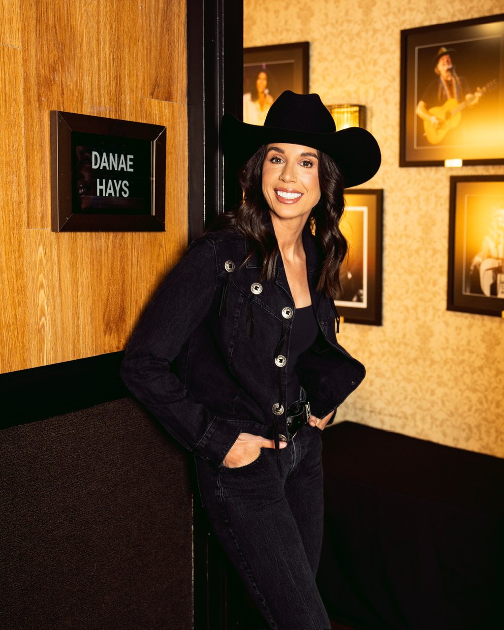 Danae Hays smiling in a black cowboy hat and denim jacket in a Ryman dressing-room doorway, next to a DANAE HAYS name sign.