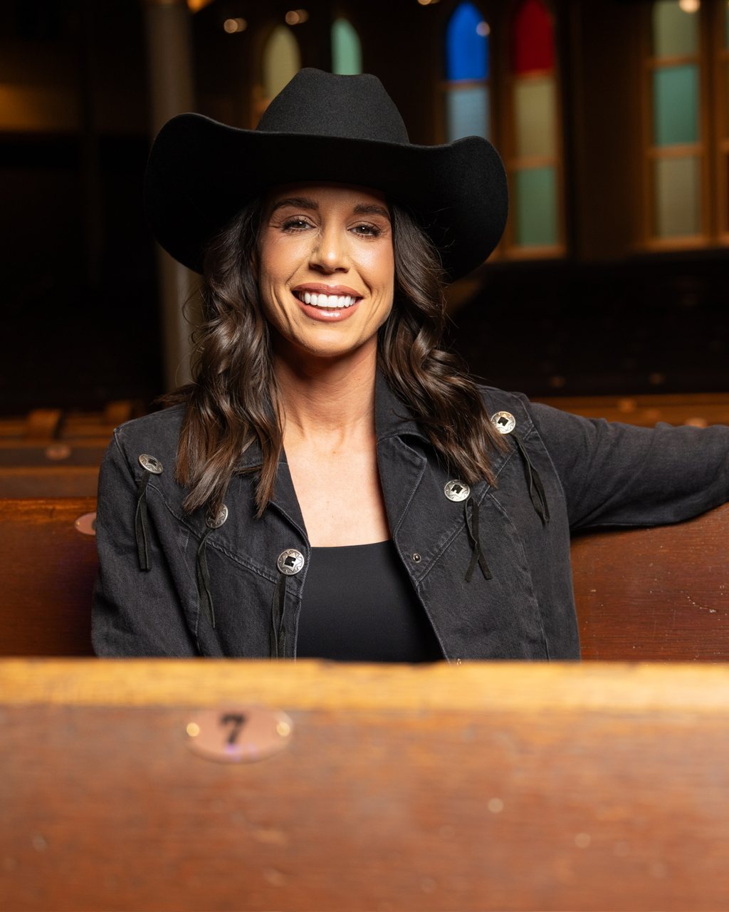 Danae Hays smiling in a black cowboy hat and denim jacket, seated in a wooden pew at the Ryman Auditorium with her arm resting along the pew in front of her.
