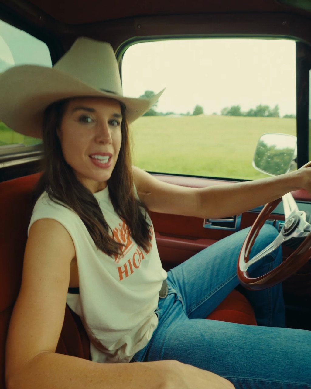 Danae Hays in a straw cowboy hat smiles at the camera from the driver's seat of a pickup with red upholstery, with a green field outside the window.